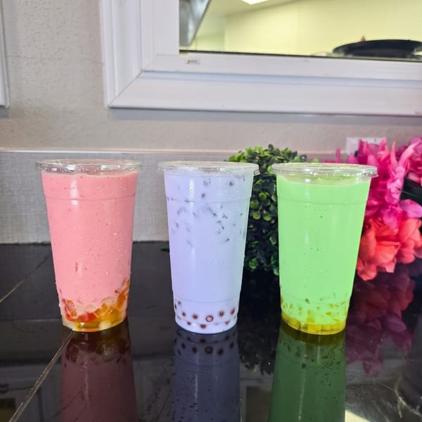 Three colorful boba tea drinks, pink, purple, and green, with toppings on a black reflective surface.