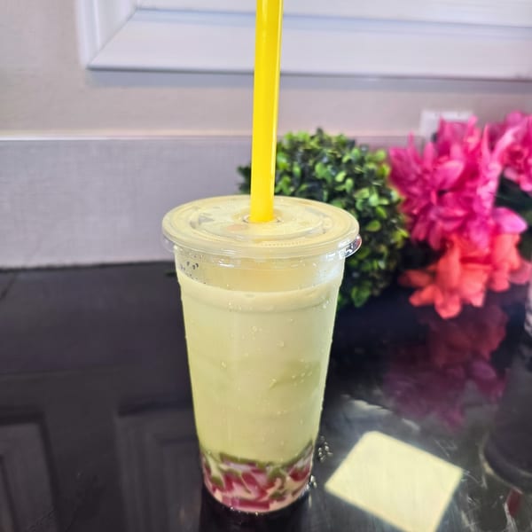 Light green boba tea with red and green jellies, a yellow straw, and pink flowers in the background.