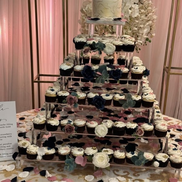 Tiered display of cupcakes with white frosting, gold accents, and decorative flowers on a table.