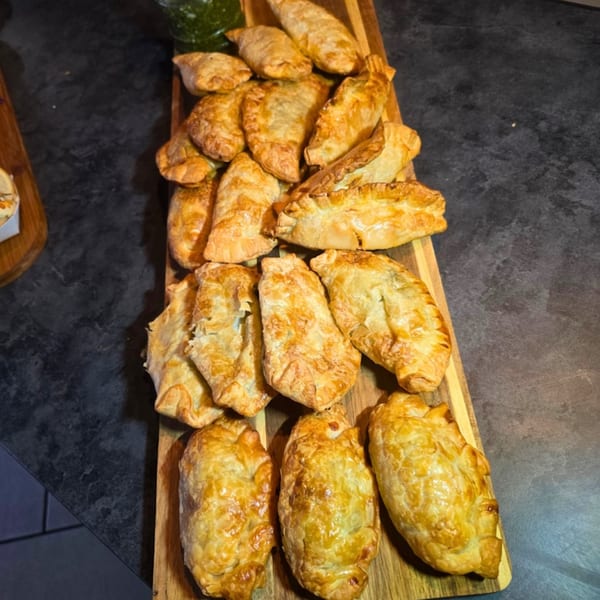 Golden-brown empanadas and pastries arranged on a wooden serving board, with a small dish of green sauce.