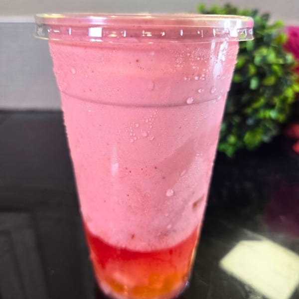 A vibrant pink and red layered drink in a clear plastic cup with condensation, against a dark background.
