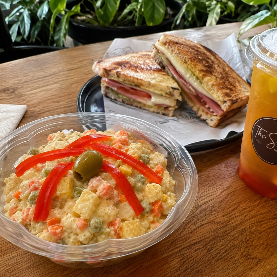 Creamy pasta salad with vegetables and olives, toasted ham and cheese sandwiches, and an orange drink on a wooden table.